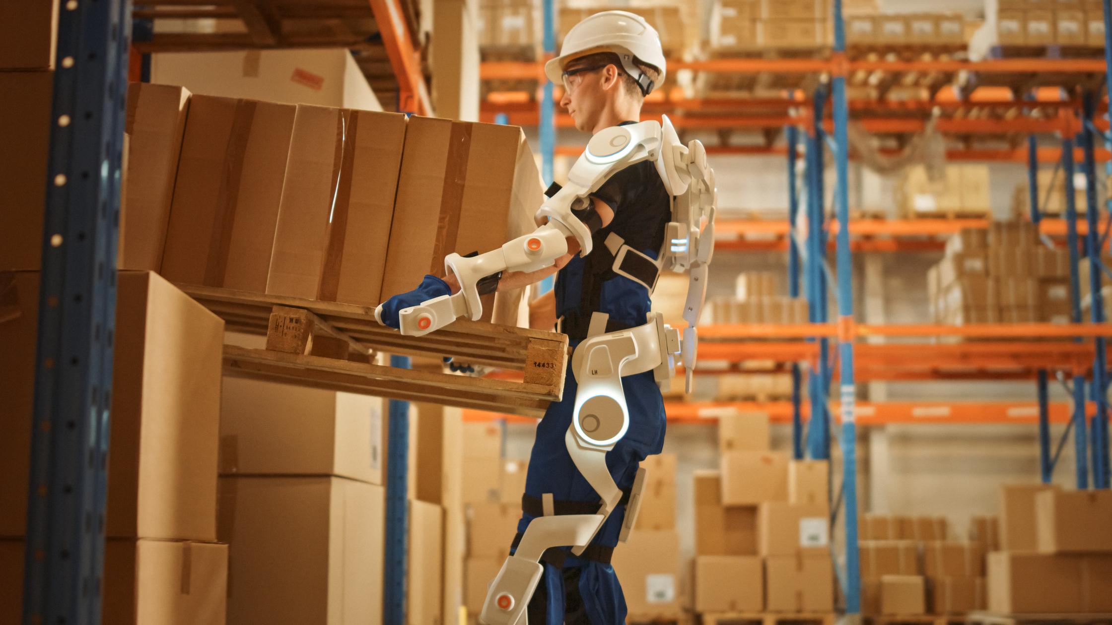 Worker wearing advanced full body powered exoskeleton, lifts heavy pallet full of cardboard boxes