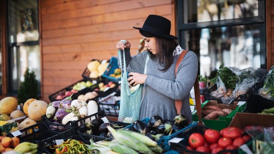A person buying fruit and vegetables at outside market, she putting vegetable in carrying bag.