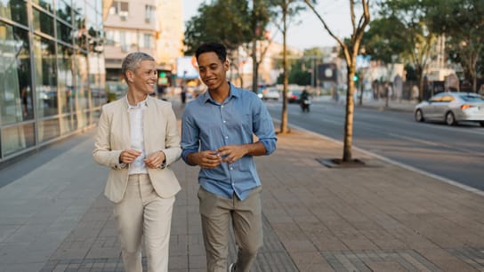 A businesswoman in a suit and a young man in casual clothing walking side by side in a city sidewalk,
