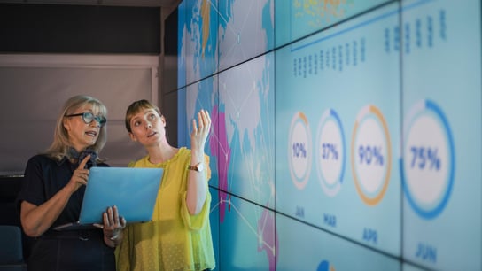 een vrouw houdt een laptop vast en bespreekt gegevens met een andere vrouwelijke collega van een interactief scherm