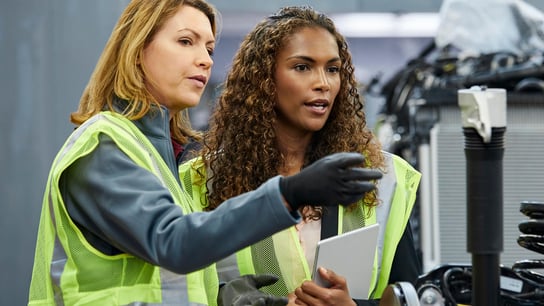 Two female engineers discussing over car chassis at automobile industry. They are wearing reflective clothing.