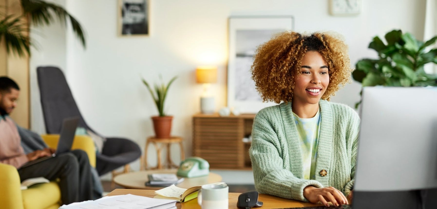 Smiling female freelancer using computer while colleague in background. Businesswoman is working at desk in home office. They are in apartment.