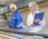 Two woman in food industry stood at conveyor belt wearing protective clothing.