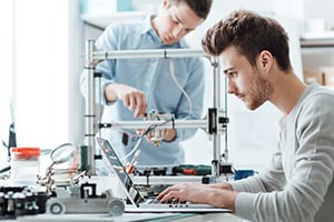 A beginner's guide to standards Two men working in a room, one is seated in front a of a laptop, and the other is in the background, working on a 3D printer