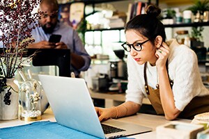 A woman works on a laptop in an office, while a man in the background looks at his mobile phone