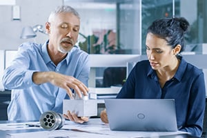 Two older people conversing about a product idea, the woman is using a laptop, and the man next to her has the product in his hands