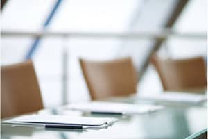 Out of focus chairs lined along a glass table with paper and pen in front of each seat.