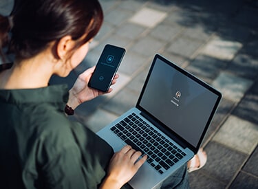 A woman sitting in front of a laptop, holding a mobile device in one hand