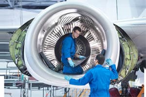 A engineer kneels next to a aeroplane engine, and looks at a tablet