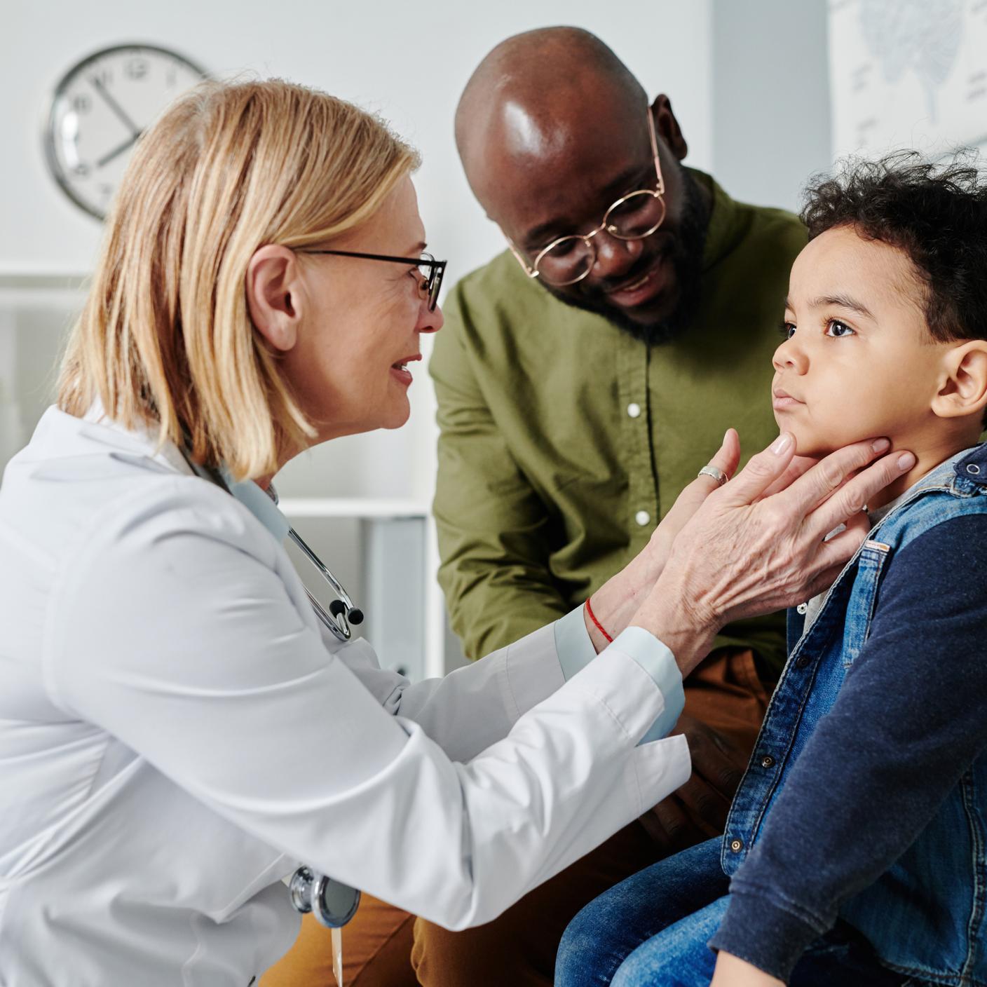 Quality in Healthcare - Female endocrinologist touching neck of little child while checking his thyroid during visit to clinics with his father