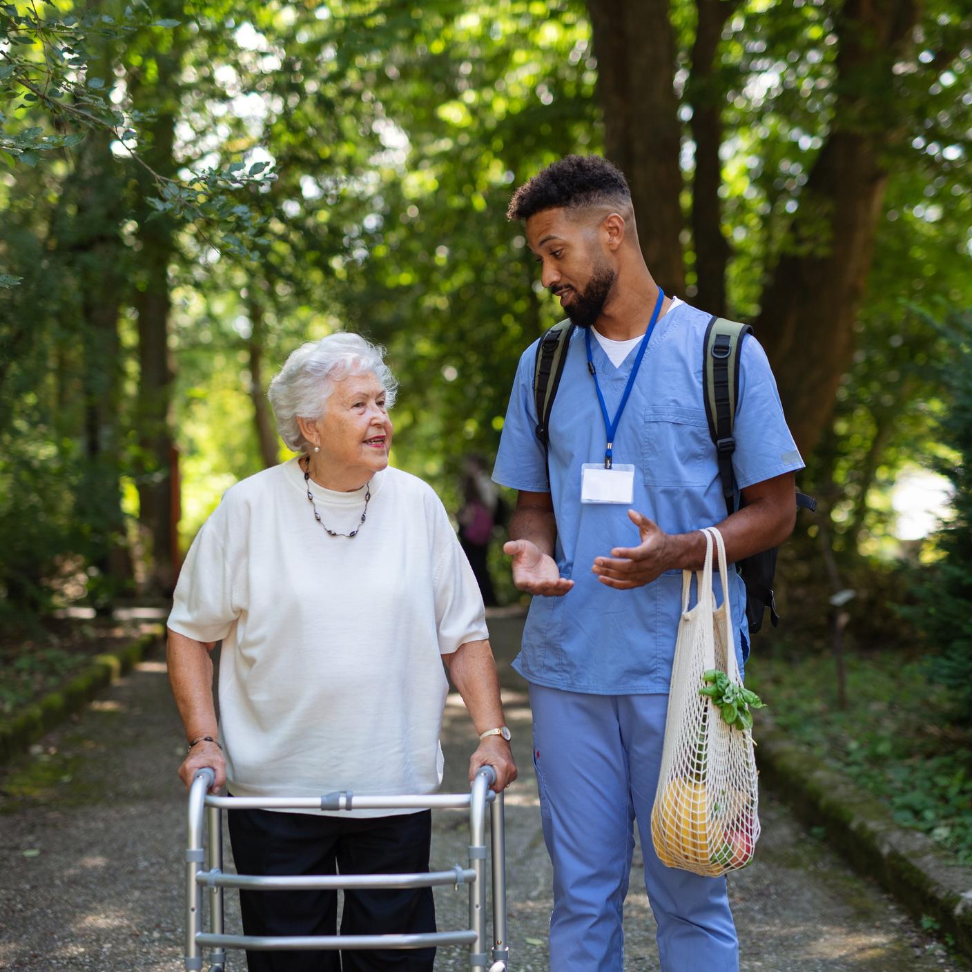 Quality in Healthcare - Caregiver man with shopping bag taking senior woman with walker for a walk to shop