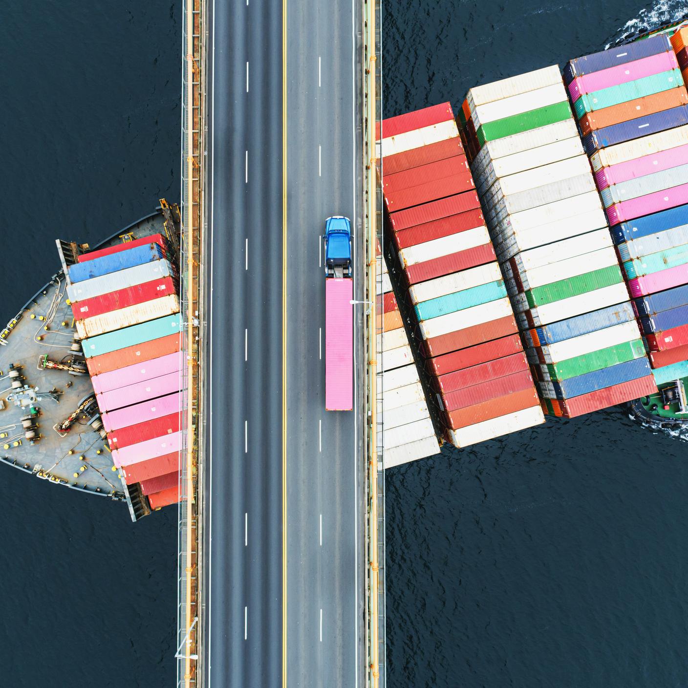 Aerial view of a container ship passing beneath a bridge while semi truck crosses above