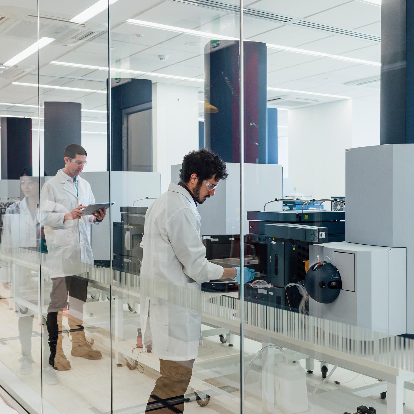 Two scientists can be seen working with large machinery through a clear glass wall in a laboratory.