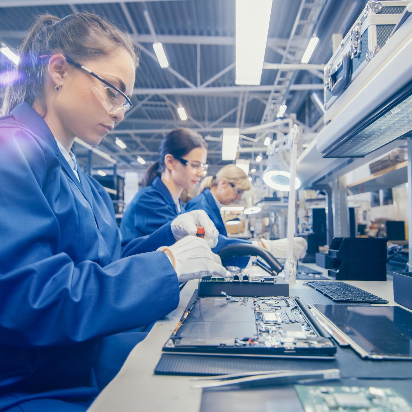 Electronics Factory Worker in Blue Work Coat and Protective Glasses is Assembling Laptop's Motherboard with a Screwdriver.