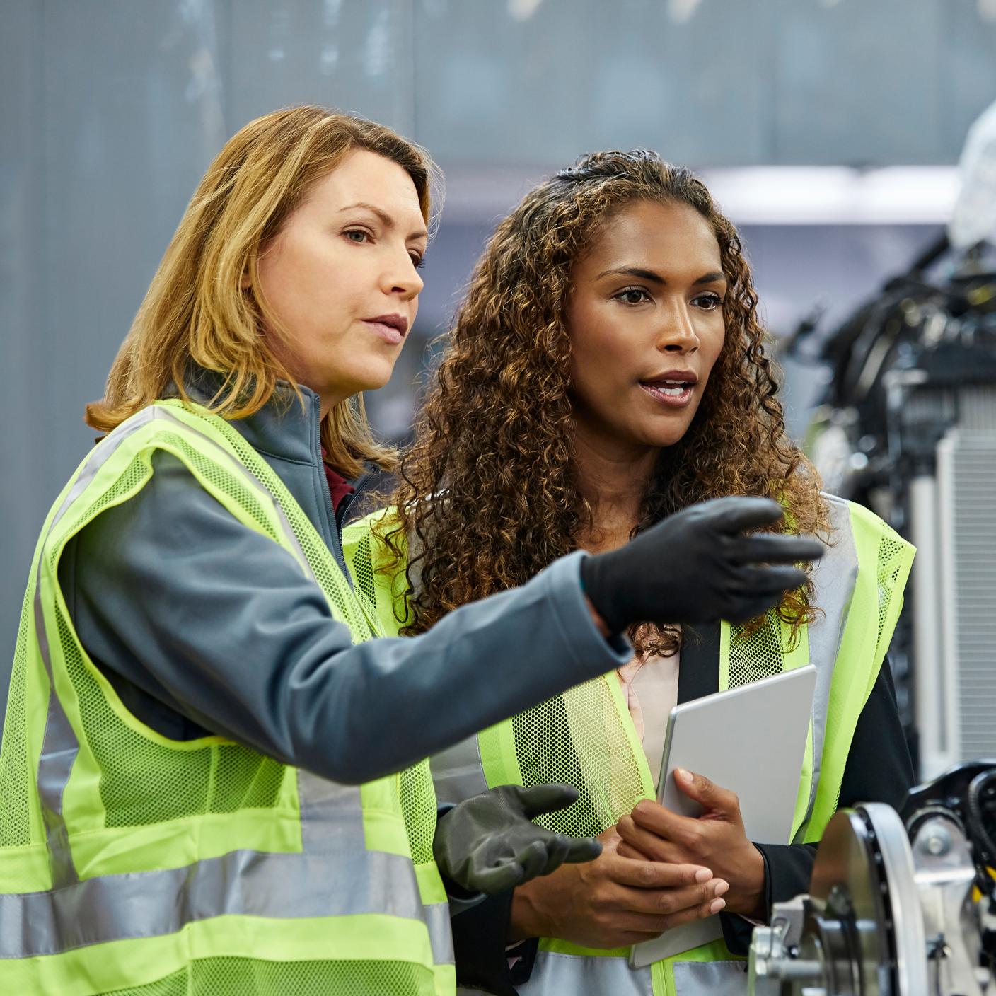 Mature female supervisor pointing to colleague by car part at factory. Confident engineers are discussing over car chassis at automobile industry. They are wearing reflective clothing. 