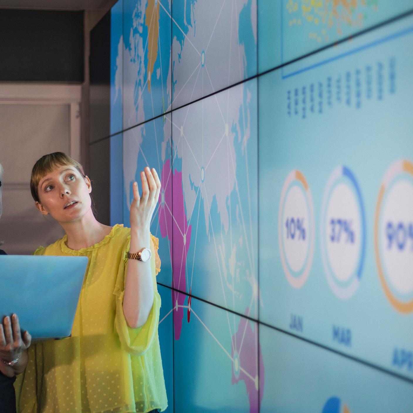 An experienced woman mentors a female colleague, the mature woman is holding a laptop as they debate data from an interactive display; they are both wearing smart casual clothing.