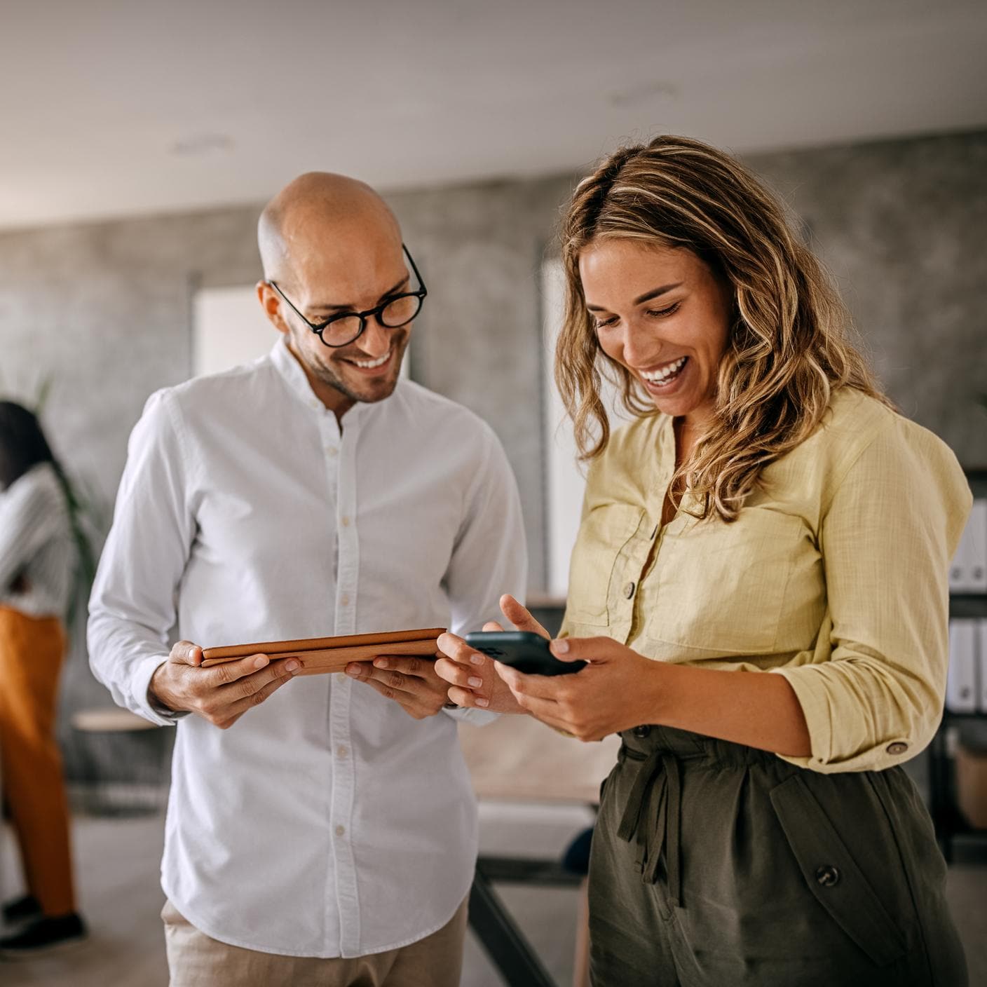 Cheerful and smiling young successful female businesswoman standing with colleague looking at smartphone in modern office and coworking space