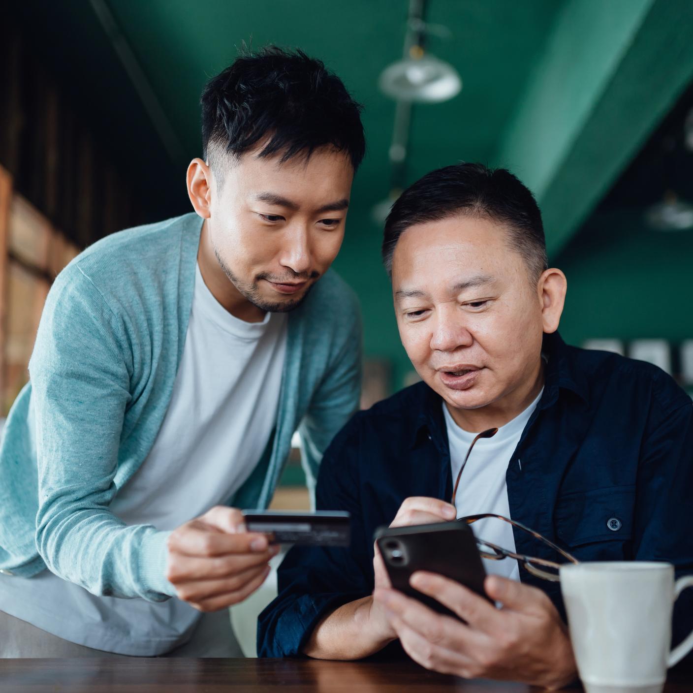 アジア系の男性とその親が、モバイルアプリでオンラインショッピングを楽しみ、自宅でクレジットカード決済をしている様子