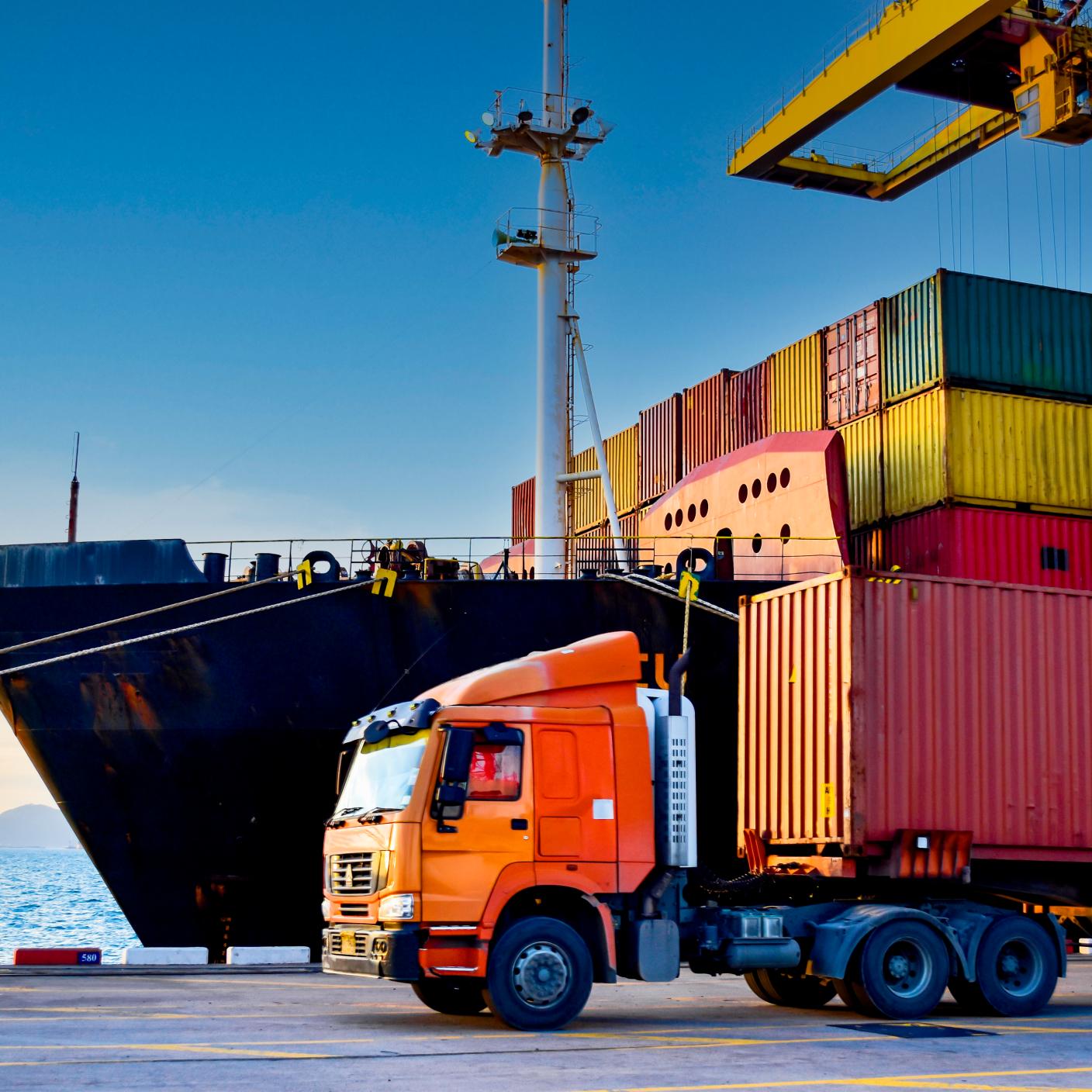 Truck carrying forty-foot container leaving port terminal with ship and quay crane on the background. Seaport operation activities, container shipping, and logistics.