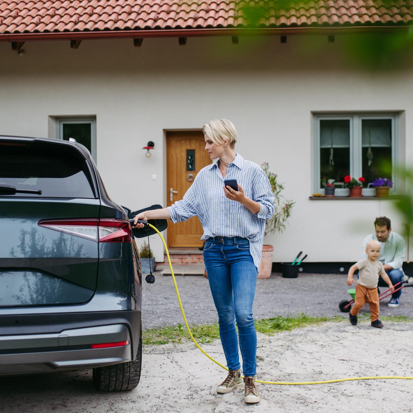 Mother charging electric car in front of house, holding smartphone. EV charging app. Electric vehicle with charger in charging port.