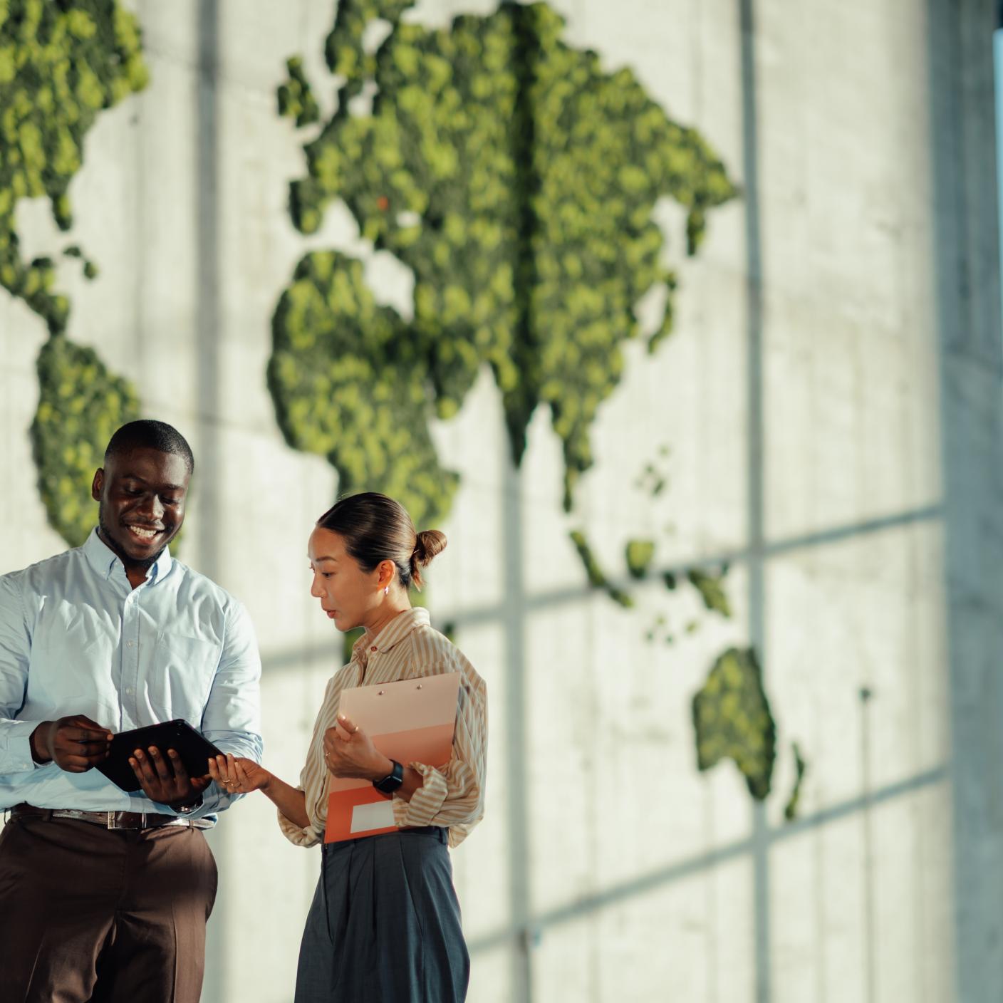 Two business colleagues are discussing global strategies in a modern office with a large green world map.