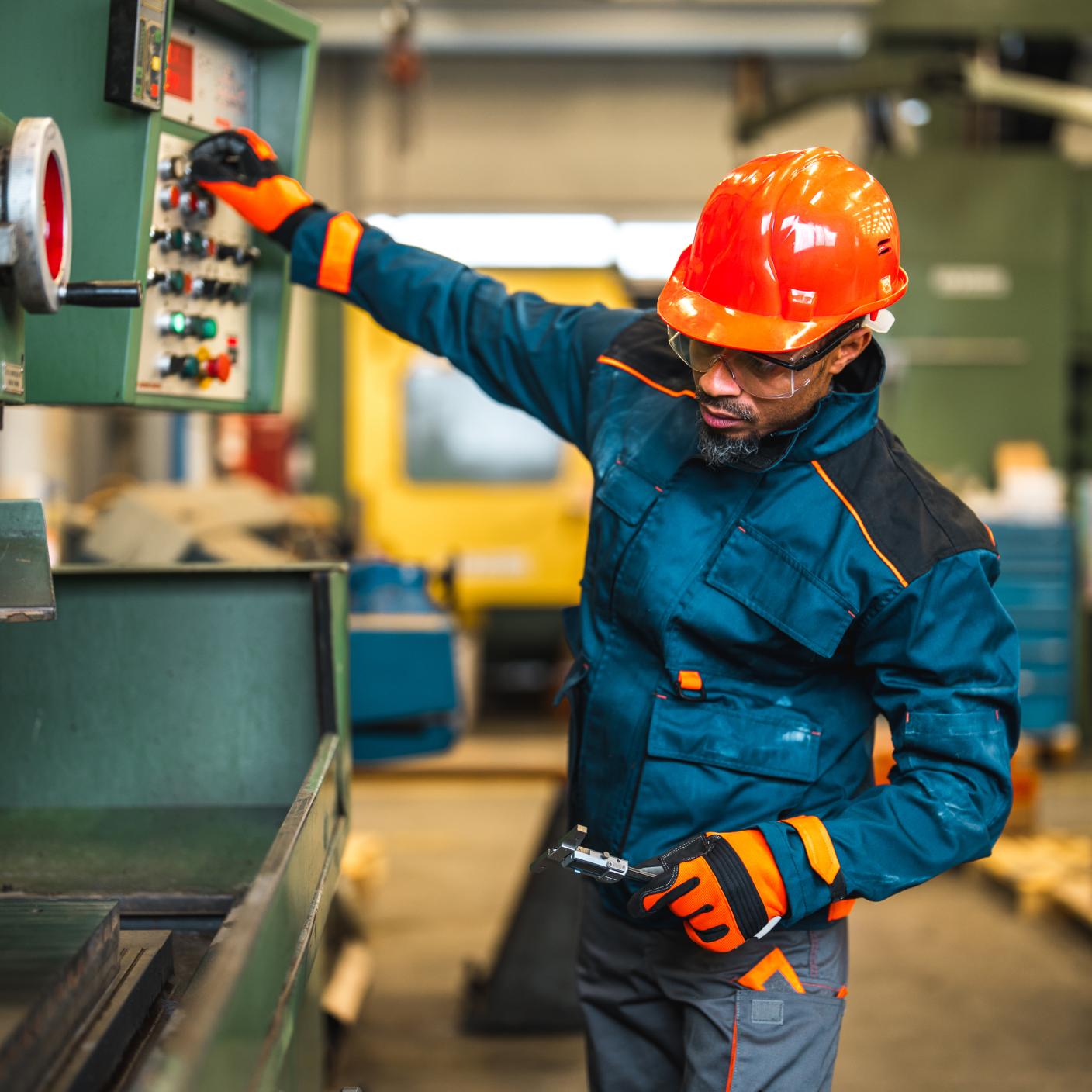 Mid adult Black blue collar worker operating an industrial machine in a factory. He is wearing protective clothes and wearing a hard hat. Pressing buttons on a machine.