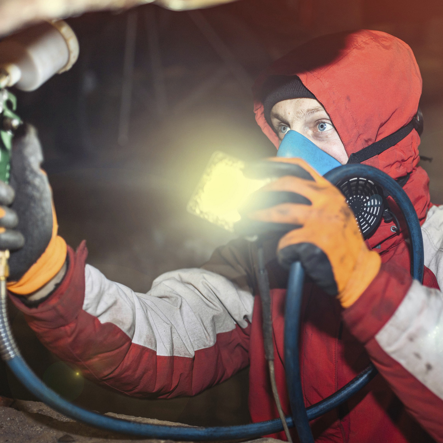 Worker in protective attire painting under a car with an orange light.