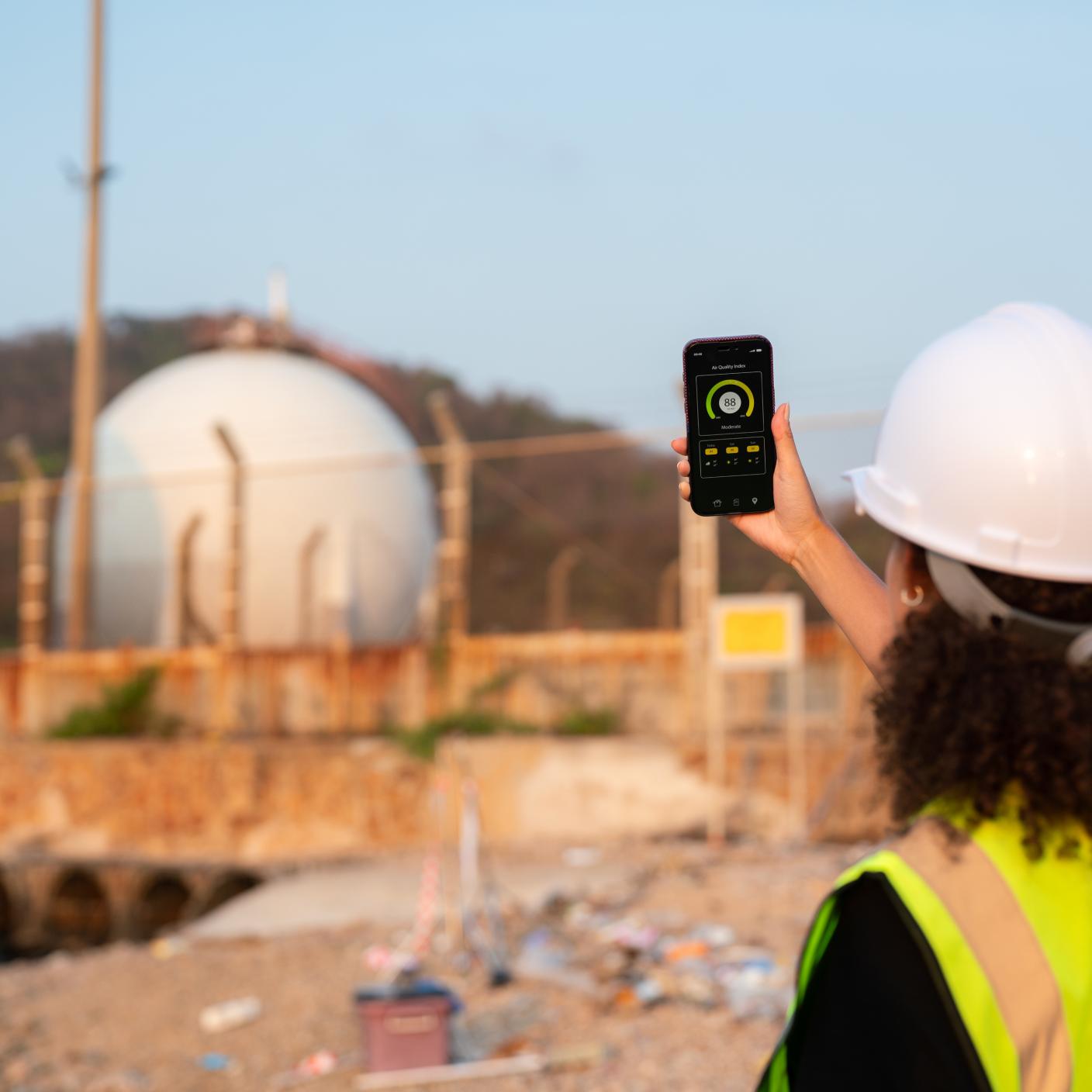 Environmental engineer checking air quality via mobile app at industrial plant. Environmental specialist wearing safety helmet and high visibility vest using smartphone application to monitor air quality levels at industrial facility, representing emission control, pollution monitoring, and environmental compliance.