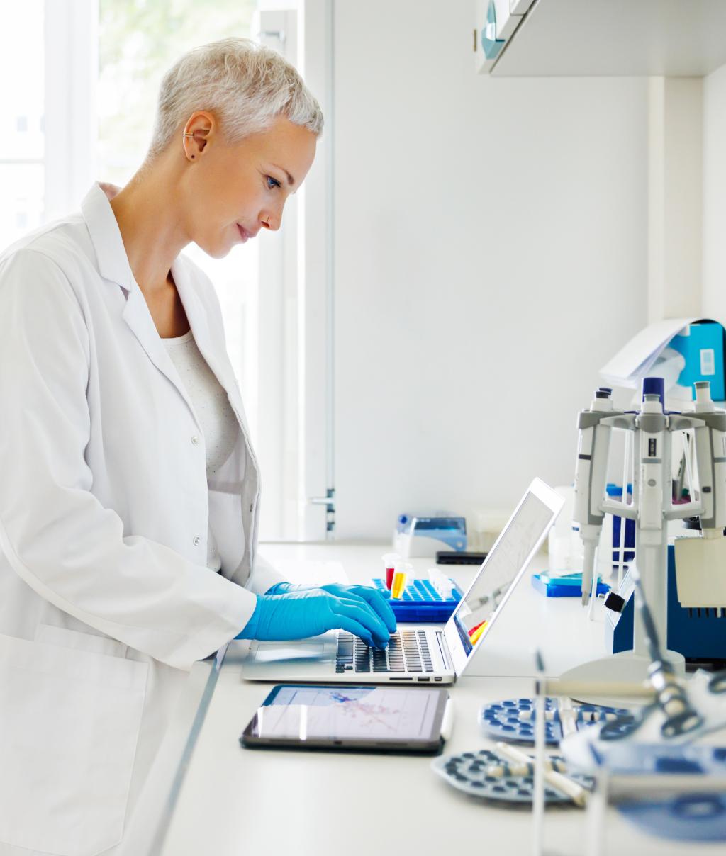 A scientist, dressed in a lab coat and gloves, focuses intently on analyzing data using a laptop and tablet. The surrounding workspace is equipped with advanced instruments, emphasizing precision and technology-driven science.