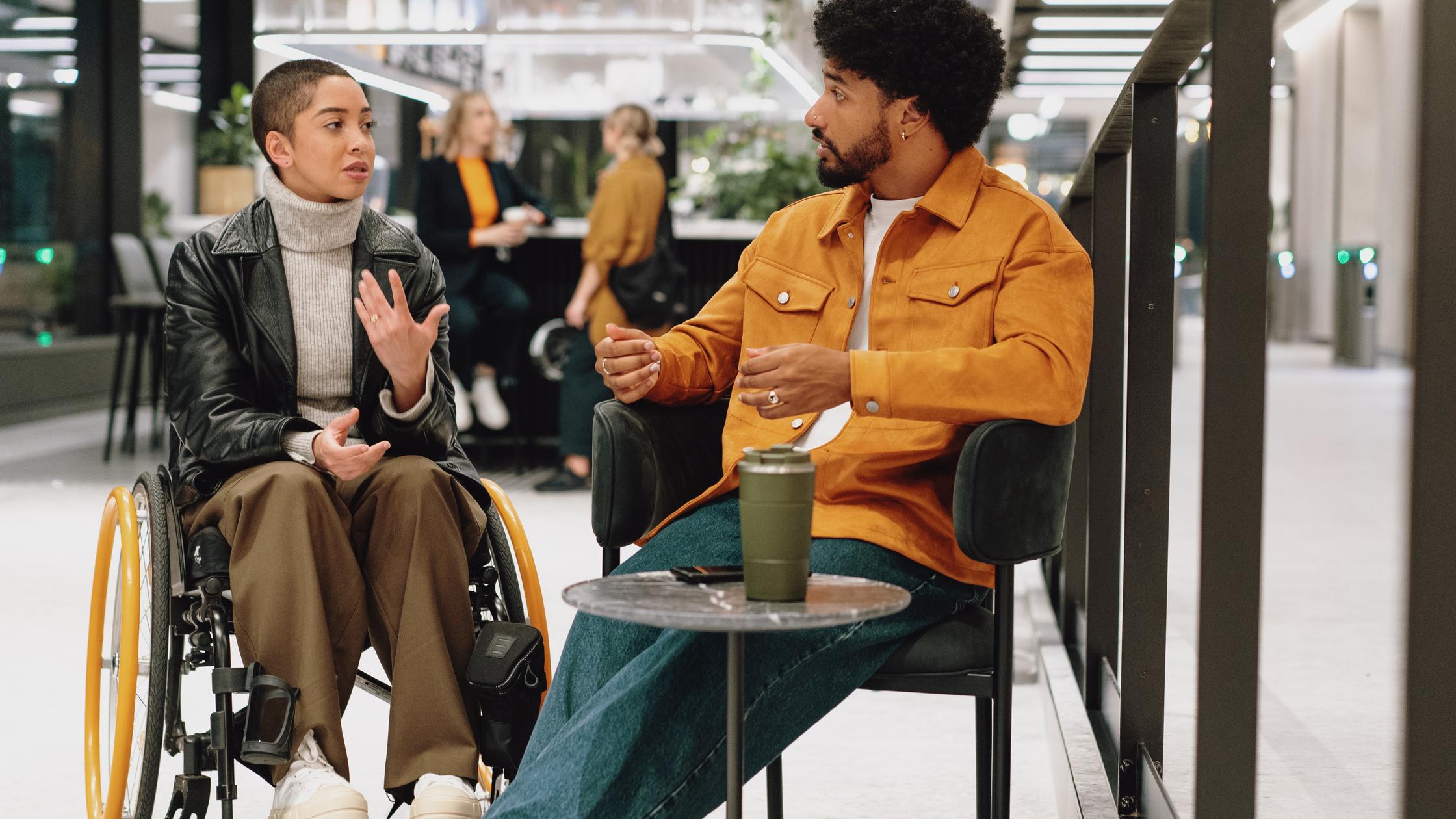 Driving business improvement - Young person in black leather jacket chats with colleague around coffee table in lobby area