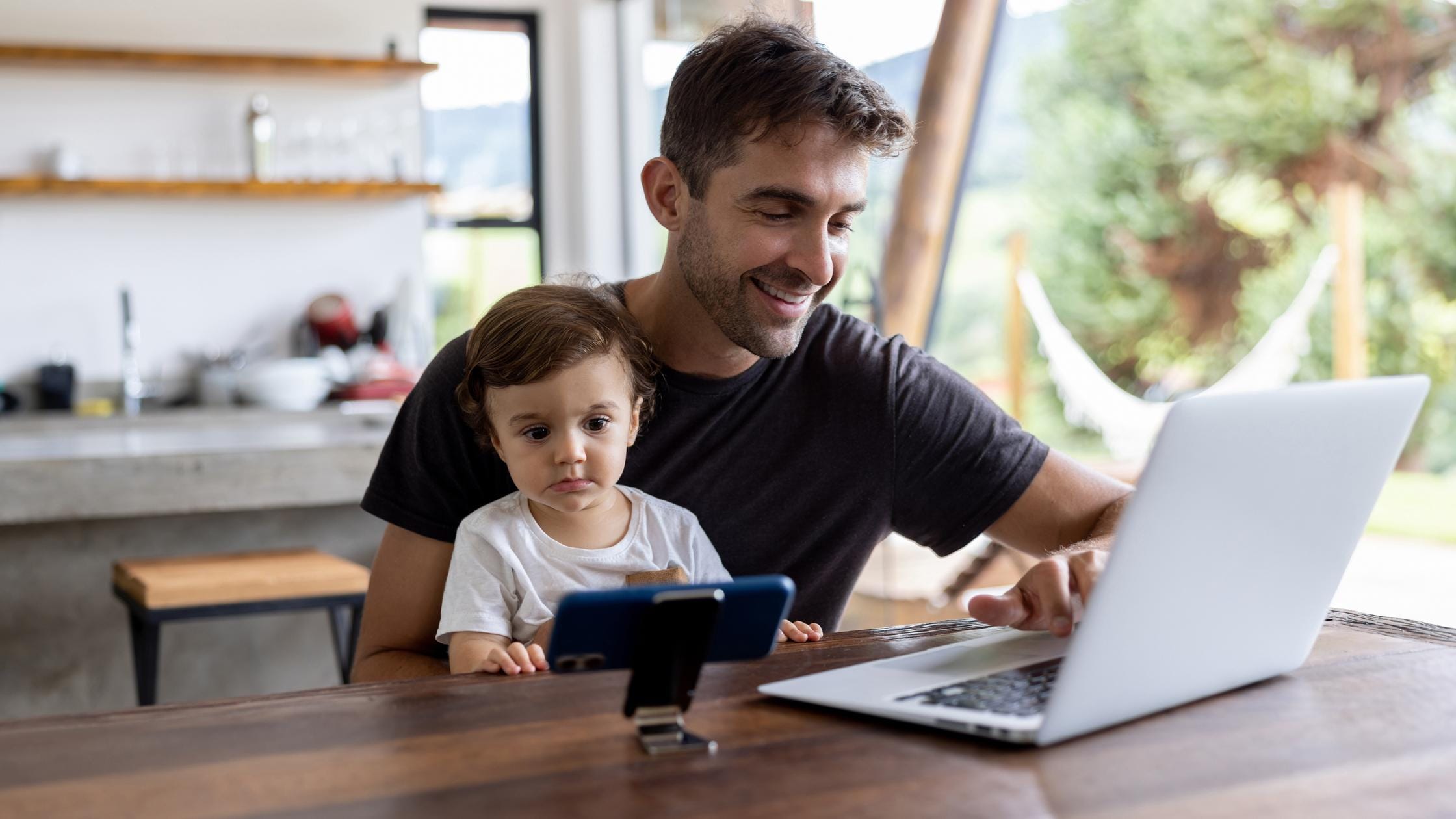 Happy father working on his laptop while his son watches tv on his cell phone