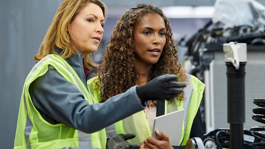 Two female engineers discussing over car chassis at automobile industry. They are wearing reflective clothing.