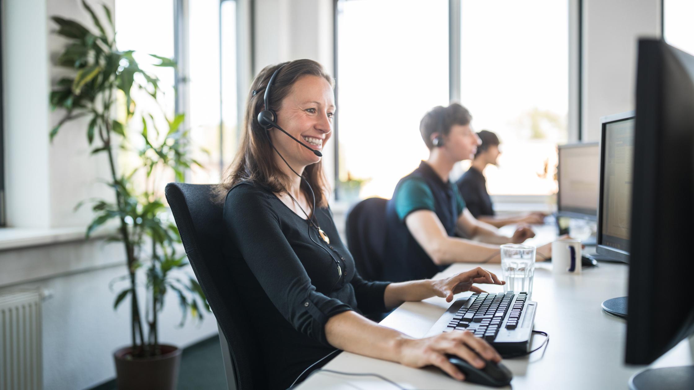 Smiling female customer service representative wearing headset using computer at desk in office