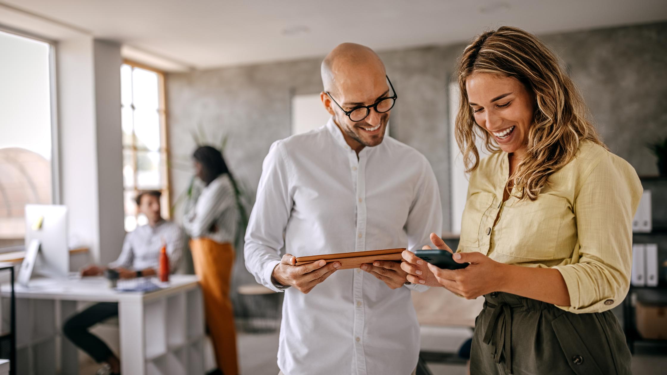 Cheerful and smiling young successful female businesswoman standing with colleague looking at smartphone in modern office and coworking space