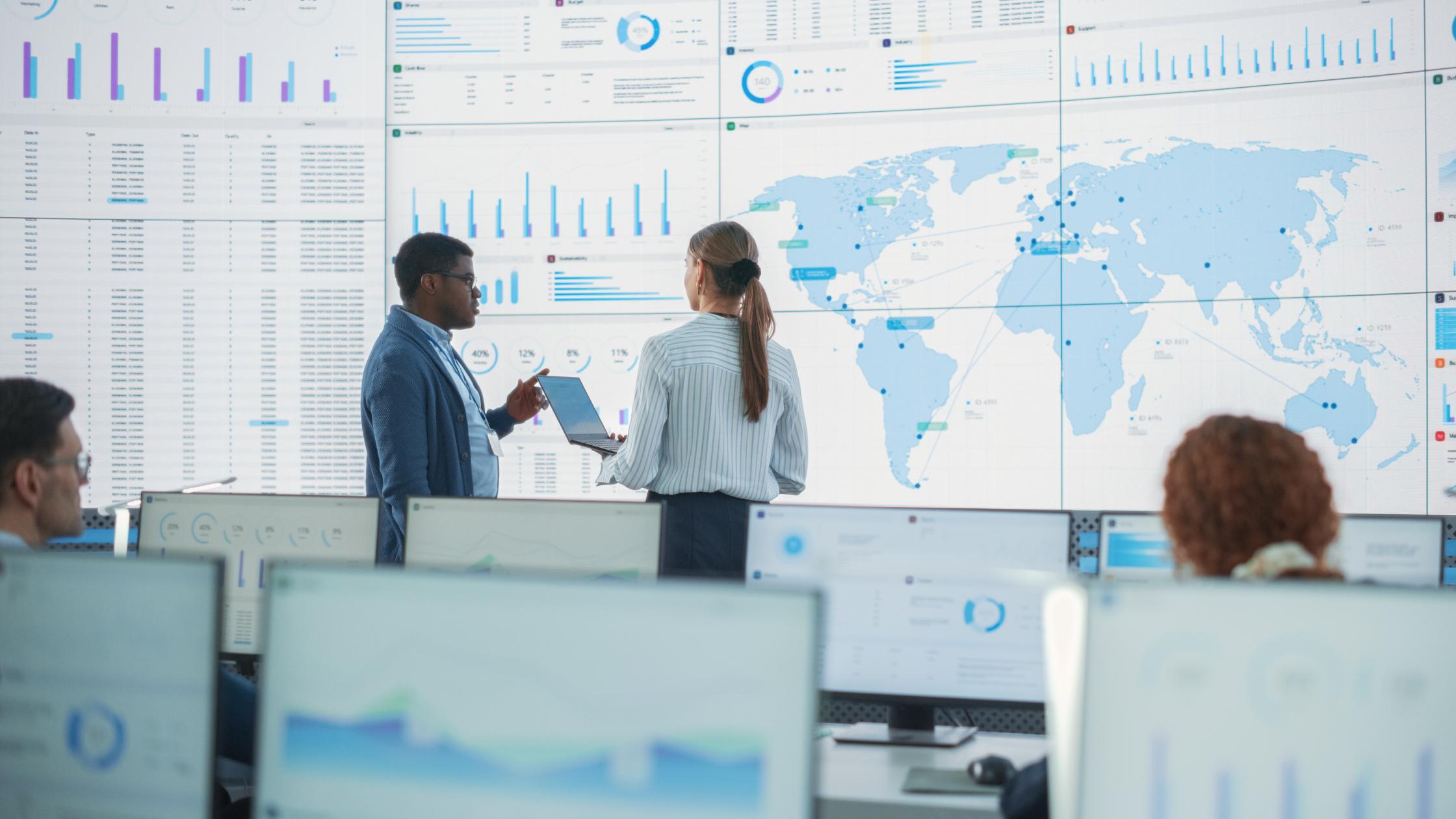 Male Analyst Talks To Caucasian Female Manager In Front Of Big Digital Screen With World Map In Monitoring Office. Diverse Employees Working Behind Desktop Computers In Logistics Company.