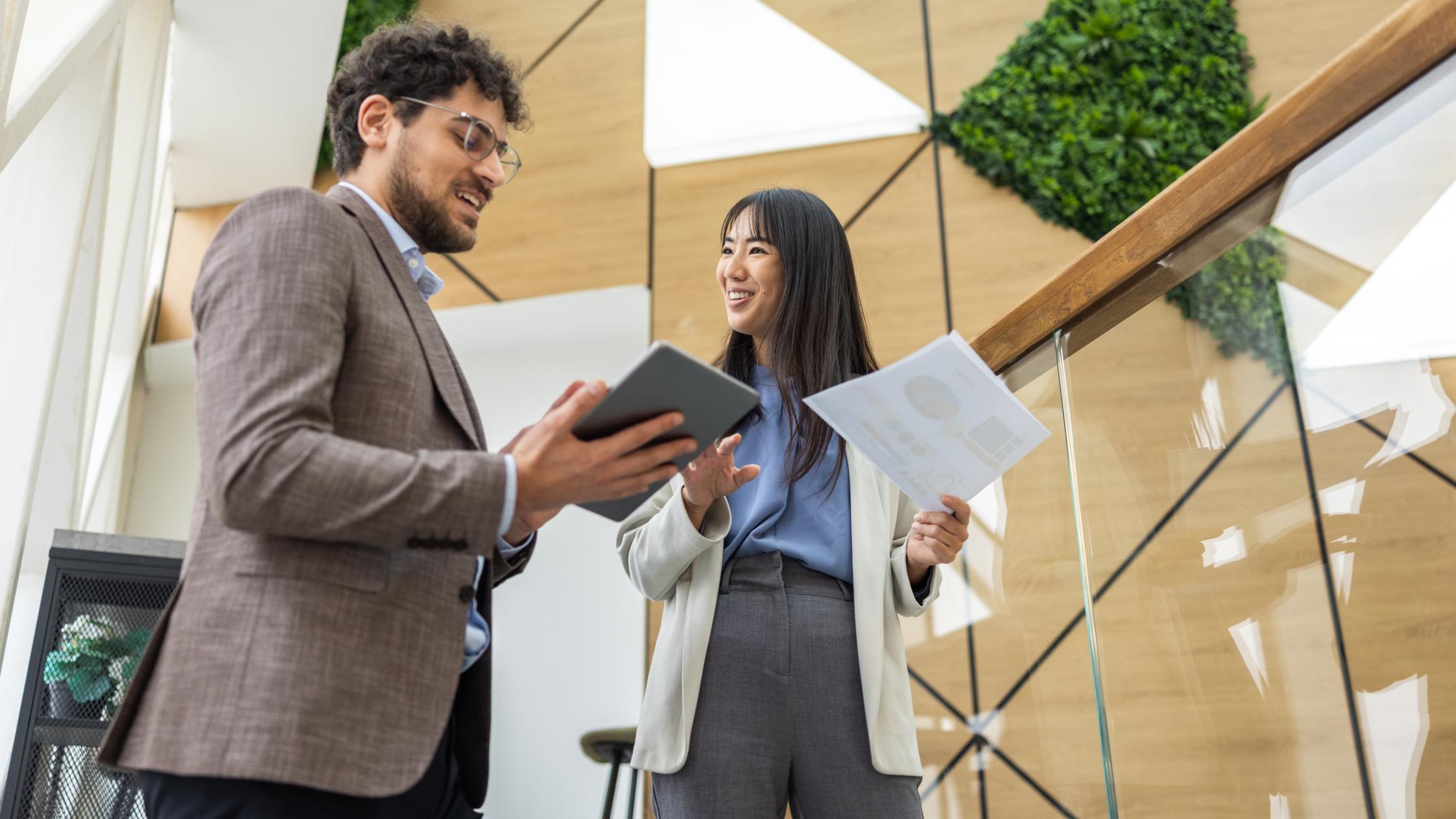 Two professionals standing together in a modern workspace, reviewing documents and discussing certification processes.