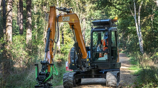 A compact excavator with a grading attachment works on a forest trail, surrounded by tall trees and dense greenery.