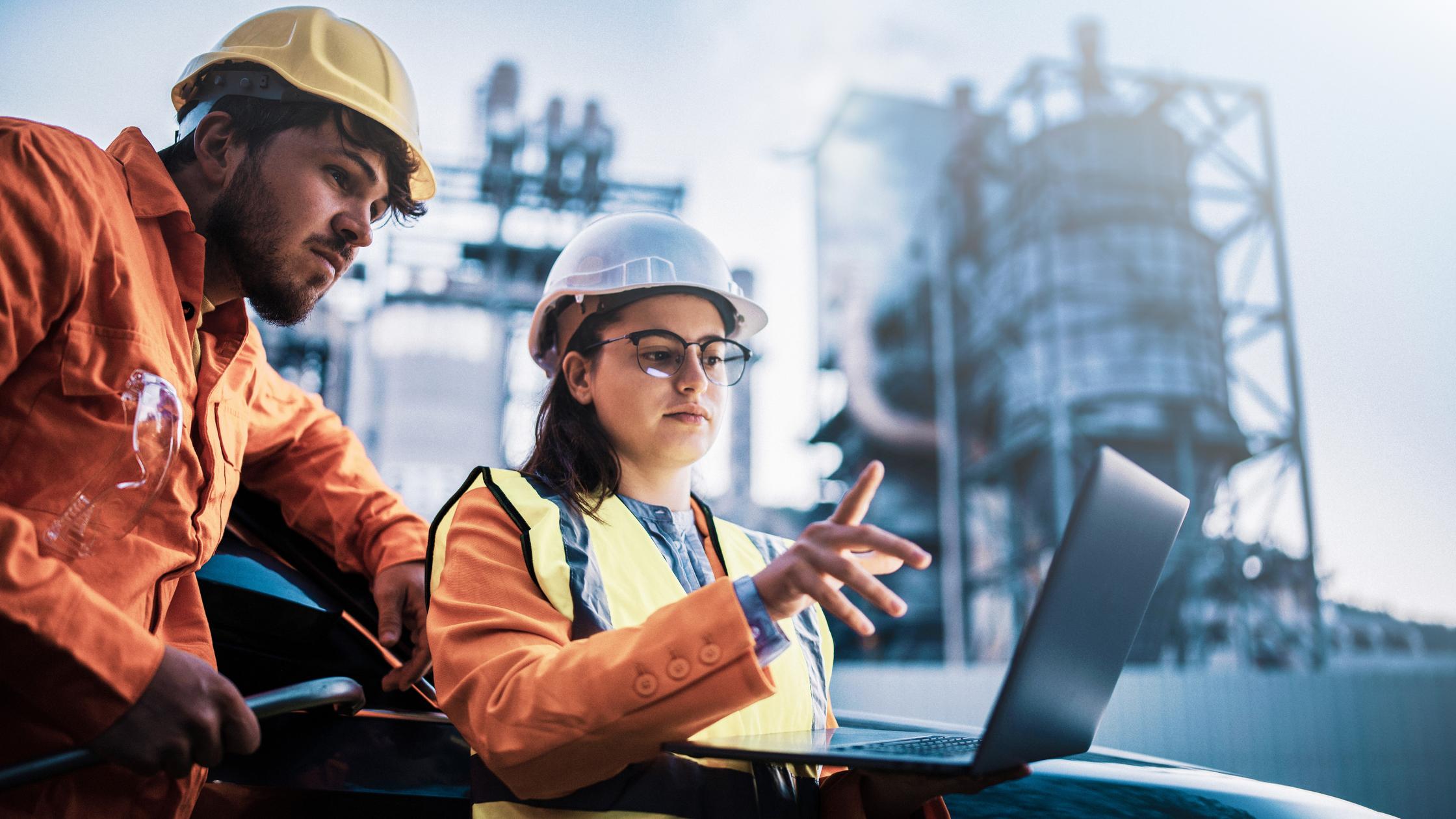 Two associates in safety attire reviewing data on a laptop in front of a power plant.