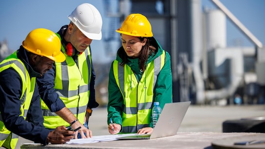 Three engineers in safety attire discussing at an industrial facility writing on paper next to a laptop.