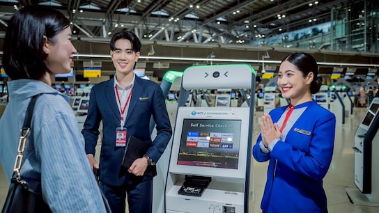 Two airport staff members, a man in a dark suit and a woman in a blue uniform, greet a passenger at an AOT Suvarnabhumi Airport self-service check-in kiosk. 
